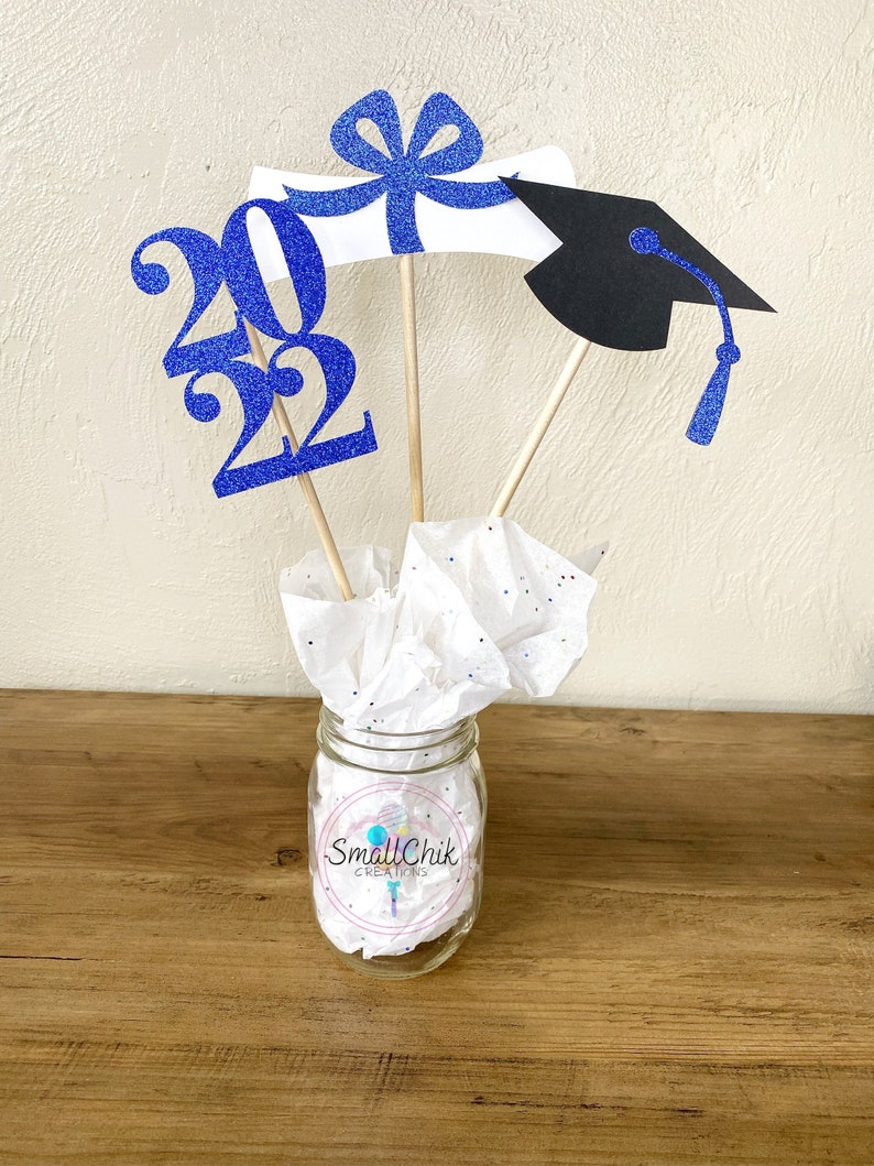 May include: Graduation centerpiece with a blue glitter 2022, a white diploma with a blue bow, and a black graduation cap with a blue tassel. The centerpiece is in a clear glass jar with white tissue paper.