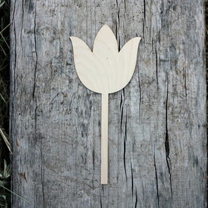 May include: A wooden cutout of a tulip flower with a long stem. The cutout is light brown and is placed on a weathered wooden surface.