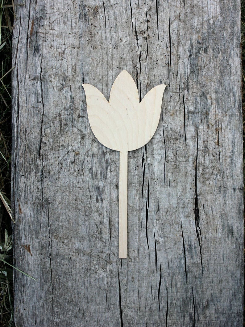 May include: A wooden cutout of a tulip flower with a long stem. The cutout is light brown and is placed on a weathered wooden surface.
