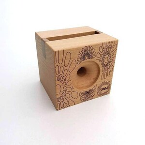 May include: A wooden phone stand with a circular cutout for a phone and a decorative pattern of circles on the front. The stand is made of light wood and has a natural finish. The text "G&M" is engraved on the front of the stand.