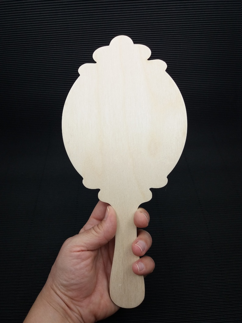 May include: A wooden hand mirror with a decorative, ornate frame and a long handle. The mirror is blank and ready to be painted or decorated.