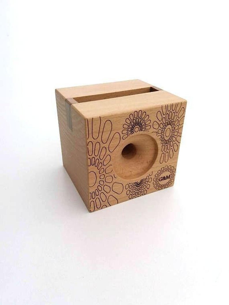 May include: A wooden phone stand with a geometric pattern and the text "G&M" on the side. The stand has a slot for holding a phone.