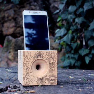 May include: A wooden phone amplifier with a circular cutout and a geometric pattern. The amplifier is made of light wood and has a dark brown finish. A white smartphone is placed in the amplifier. The amplifier is sitting on a wooden surface.