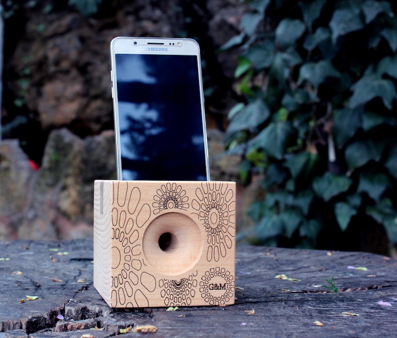 May include: A wooden phone amplifier with a circular cutout and a geometric pattern. The amplifier is made of light wood and has a dark brown finish. A white smartphone is placed in the amplifier. The amplifier is sitting on a wooden surface.