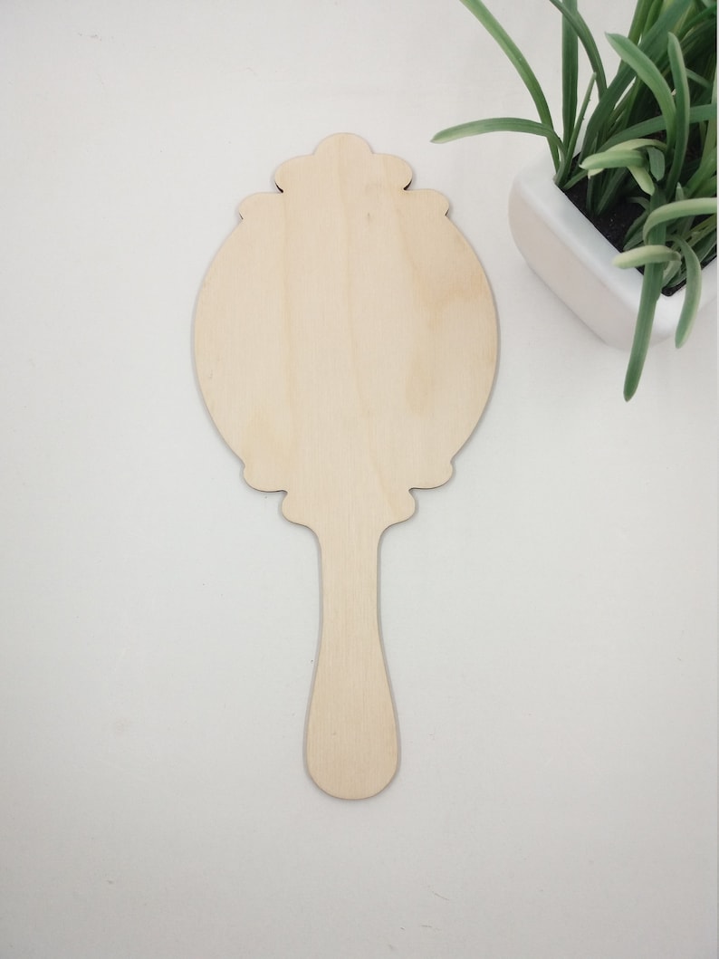 May include: A wooden hand mirror with a decorative, ornate frame and a long handle. The mirror is unfinished and ready to be painted or stained.