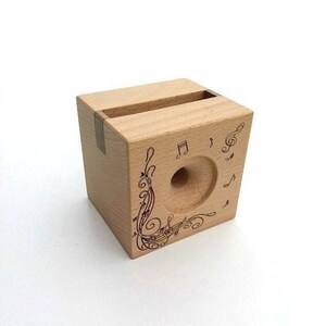 May include: A wooden phone stand with a cutout for a phone and a design of musical notes and treble clefs. The stand is made of light wood and has a natural finish.