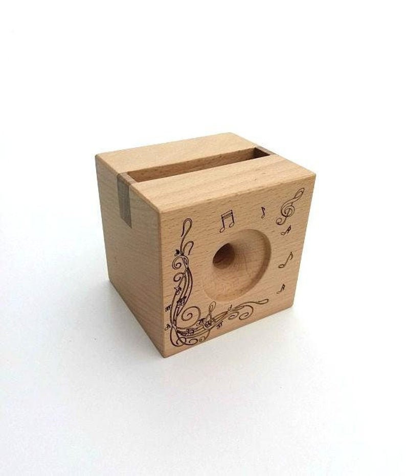 May include: A wooden phone stand with a cutout for a phone and a design of musical notes and treble clefs. The stand is made of light wood and has a natural finish.