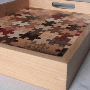 K&ouml;nnte beinhalten: Ein Holztablett mit Griff, das mit einem Mosaik aus Puzzle-Teilen aus verschiedenen Holzarten verziert ist.
