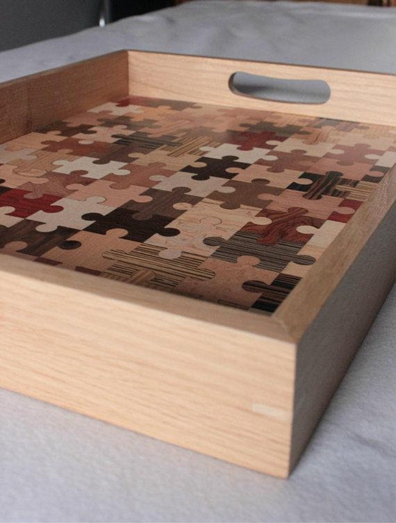 K&ouml;nnte beinhalten: Ein Holztablett mit Griff, das mit einem Mosaik aus Puzzle-Teilen aus verschiedenen Holzarten verziert ist.