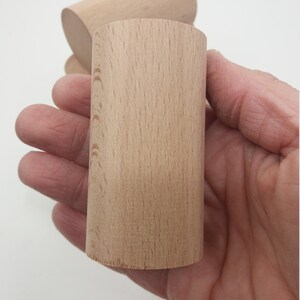 K&ouml;nnte beinhalten: Zwei unfertige Holzzylinder, einer ist teilweise hinter dem anderen sichtbar, mit einem nat&uuml;rlichen Holzmaserungsmuster. Die Zylinder werden in einer Hand gehalten.