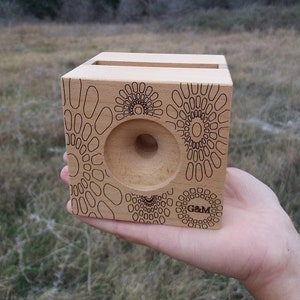 May include: A wooden phone stand with a geometric pattern and the text "G&M" engraved on the front. The stand has a cutout for a phone and a small hole in the center.