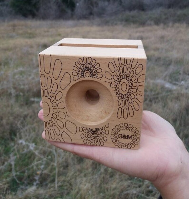 May include: A wooden phone stand with a geometric pattern and the text "G&M" engraved on the front. The stand has a cutout for a phone and a small hole in the center.