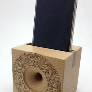 May include: A wooden phone stand with a decorative laser-etched design. The stand has a cutout for a phone and a hole for sound amplification. A Samsung phone is in the stand.