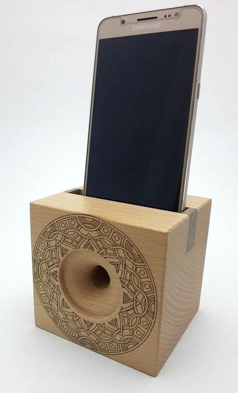 May include: A wooden phone stand with a decorative laser-etched design. The stand has a cutout for a phone and a hole for sound amplification. A Samsung phone is in the stand.
