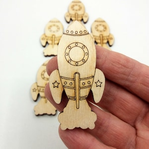 May include: A set of five unfinished wooden rocket ship cutouts. The rockets are approximately 2 inches tall and have a simple design with a round window, a pointed nose, and a fin on each side. The rockets are perfect for crafting and decorating.