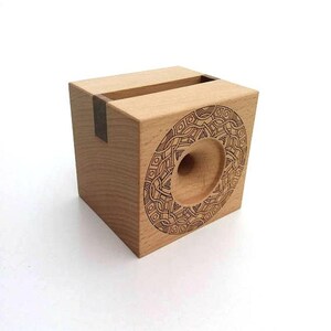 May include: A wooden phone stand with a geometric design carved into the front. The stand is made of light wood with a dark wood accent.