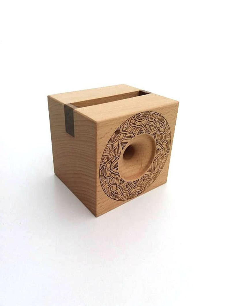 May include: A wooden phone stand with a geometric design carved into the front. The stand is made of light wood with a dark wood accent.