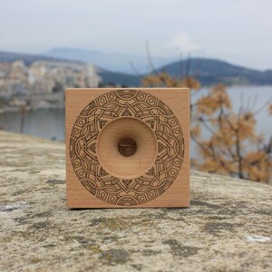 May include: A wooden square with a circular design engraved on the front. The design is a mandala with a central hole. The background is a blurry image of a city and a body of water.