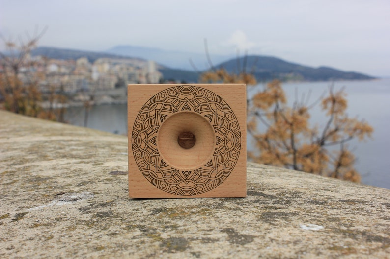 May include: A wooden square with a circular design engraved on the front. The design is a mandala with a central hole. The background is a blurry image of a city and a body of water.
