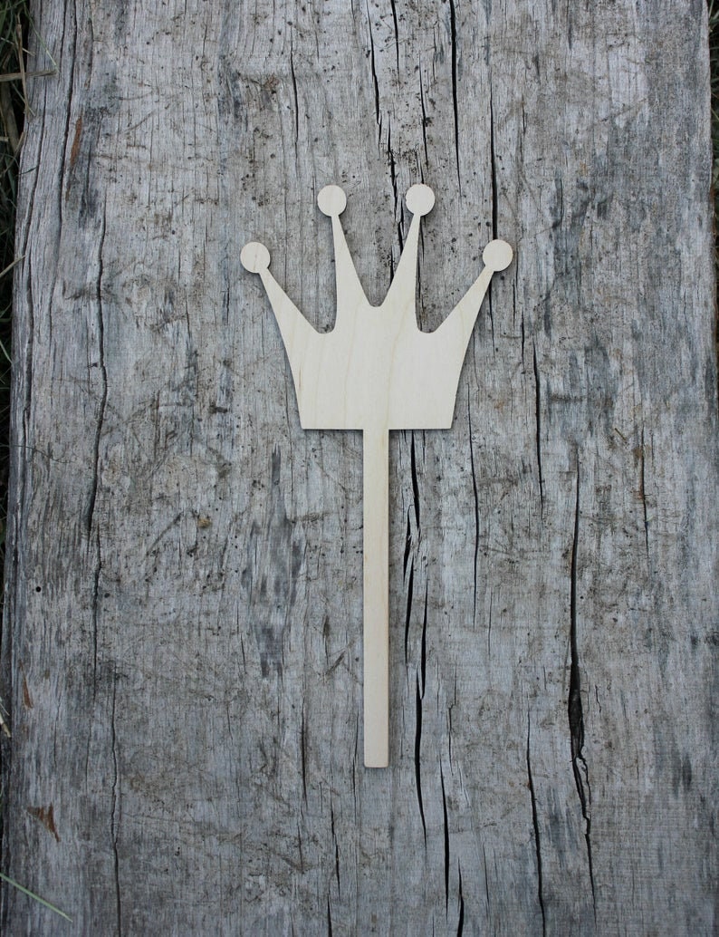 May include: A wooden crown cutout with a stick attached to the bottom. The crown is light brown and has five points.