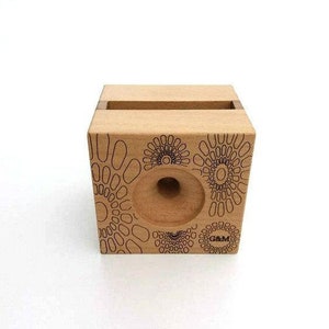 May include: A wooden phone stand with a geometric pattern and the text "G&M" printed on the front. The stand has a cutout for a phone and a hole in the back for charging cables.