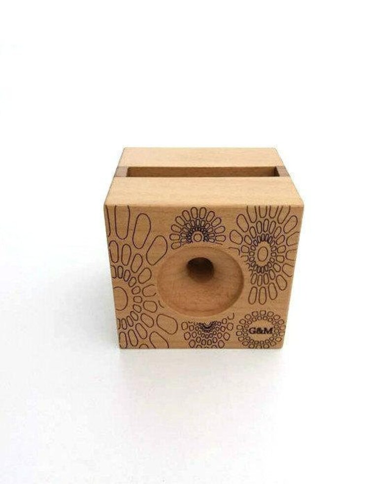 May include: A wooden phone stand with a geometric pattern and the text "G&M" printed on the front. The stand has a cutout for a phone and a hole in the back for charging cables.