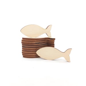 Puede incluir: Recortes de madera con forma de pez, perfectos para manualidades. La imagen muestra dos peces de color claro, uno descansando sobre una pila de piezas de madera marrón. Los peces tienen un diseño simple y limpio, ideal para varios proyectos de bricolaje. El fondo es blanco liso.