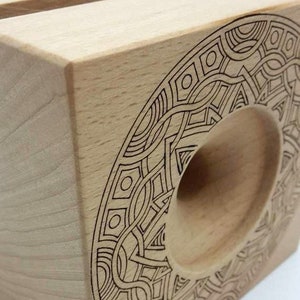 May include: A wooden phone stand with a geometric design carved into the side. The stand is made of light wood and has a circular cutout for holding a phone.