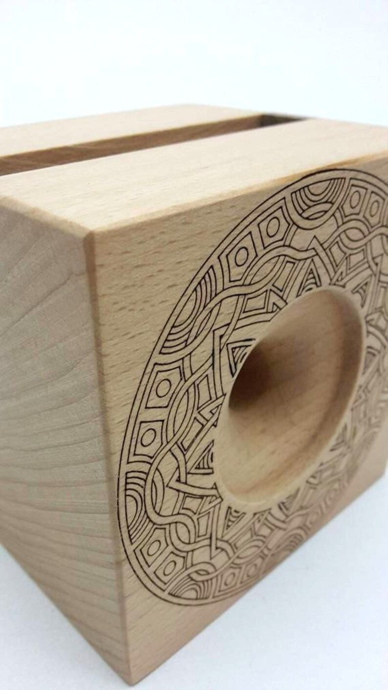 May include: A wooden phone stand with a geometric design carved into the side. The stand is made of light wood and has a circular cutout for holding a phone.