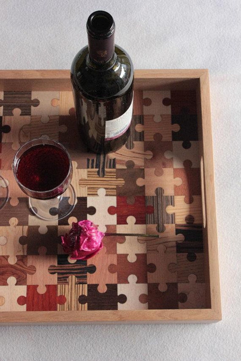 K&ouml;nnte beinhalten: Ein Holztablett mit einem Puzzle-Design aus verschiedenen Holzst&uuml;cken in verschiedenen Farben. Auf dem Tablett stehen eine Flasche Rotwein und ein Glas Rotwein sowie eine rosa Blume.