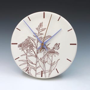 May include: A round, off-white ceramic clock with a floral and butterfly design. The clock face features brown line art of flowers and a butterfly. It has brown hour markers, a brown minute hand, and a light blue hour and second hand.