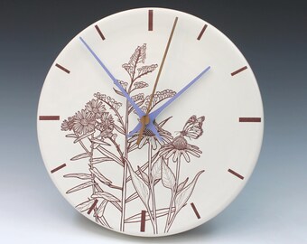 May include: A round, off-white ceramic clock with a floral and butterfly design. The clock face features brown line art of flowers and a butterfly. It has brown hour markers, a brown minute hand, and a light blue hour and second hand.