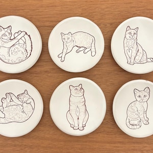 May include: Six white ceramic coasters with black line drawings of cats in various poses. The cats are depicted lying down, sitting, and stretching. The coasters are arranged on a wooden surface.