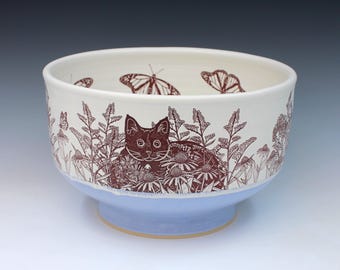May include: A ceramic bowl with a cream-colored interior and exterior featuring a dark brown line art illustration of a cat sitting among flowers and butterflies. The lower portion of the bowl is finished in a solid light blue glaze.