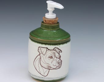 May include: A ceramic soap dispenser with a white body and a green glazed top. The front features a brown line drawing of a dog head. It includes a cork stopper at the neck and a white plastic pump mechanism at the top for dispensing liquid soap.