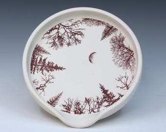May include: A white ceramic spoon rest with a forest and crescent moon design. The rim is round with a small spout. The forest design is in a dark reddish-brown color, with trees and a crescent moon in the center.