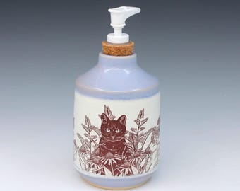 May include: A ceramic soap dispenser with a light purple glaze on the top and a cream-colored base. The body features a dark brown line art illustration of a cat surrounded by flowers. It includes a cork stopper and a white plastic pump mechanism.
