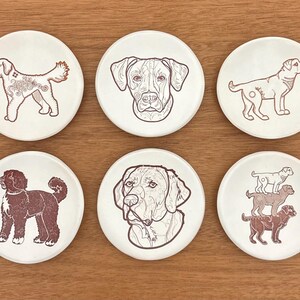 May include: Set of six ceramic coasters featuring dog illustrations in brown and white. Each coaster displays a different dog breed or a stack of dogs. The coasters are round and set on a wooden surface.