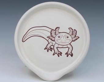 May include: A round, off-white ceramic spoon rest featuring a dark brown line art illustration of an axolotl centered on the surface. The creature is depicted with its signature feathery external gills, four legs, and a long, curved tail.