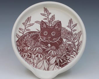 May include: A round white ceramic spoon rest featuring a detailed dark maroon illustration of a cat sitting among various wildflowers and tall grasses. The design covers the center of the dish, which has a curved indentation at the bottom edge.