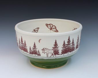 May include: A white ceramic bowl with a green base, featuring a forest scene with trees, butterflies, and a moose in a brown color. The bowl has a wide rim and is suitable for serving.