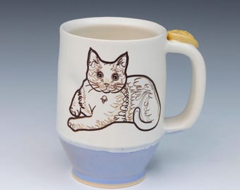 May include: A ceramic mug with a white upper body and a light blue base. The front features a dark brown line drawing of a cat in a resting position. The handle is white and includes a small, yellow, sculpted detail at the top attachment point.