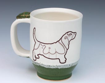 May include: A ceramic mug with a cream-colored upper body and a dark green base. The mug features a brown line-art illustration of a basset hound on the side and a small, sculpted green frog figure attached to the top of the handle.