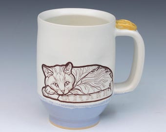 May include: A ceramic mug with a white upper body and a light blue base. A brown line drawing of a resting cat is featured on the front. The handle is white and topped with a small, yellow, circular decorative element resembling a cookie.
