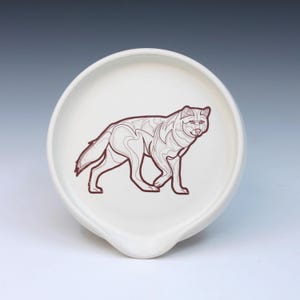 May include: A white ceramic spoon rest with a brown outline drawing of a wolf. The wolf is depicted in a stylized, geometric design. The spoon rest has a curved shape with a small lip for holding utensils.