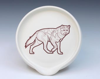 May include: A white ceramic spoon rest with a brown outline drawing of a wolf. The wolf is depicted in a stylized, geometric design. The spoon rest has a curved shape with a small lip for holding utensils.