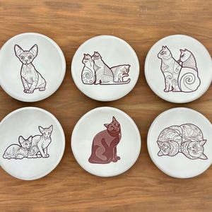 May include: Six white ceramic coasters with cat illustrations in shades of brown. Each coaster features a different cat design, including a Sphynx cat, cats cuddling, and cats in various poses. The coasters are arranged on a wooden surface.