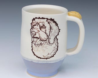May include: A ceramic mug with a white upper body and a light blue base. The front features a dark brown line drawing of a shaggy dog. The handle is white and includes a small, yellow, textured decorative accent at the top where it meets the rim.