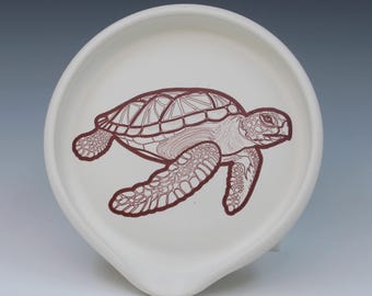 May include: A round, off-white ceramic spoon rest featuring a detailed, dark brown line drawing of a sea turtle centered on the surface. The spoon rest has a smooth, matte finish and a distinct indentation at the bottom edge for a spoon handle.
