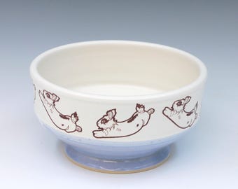 May include: A ceramic bowl with a smooth cream-colored interior and exterior. The exterior features a repeating pattern of brown line-art illustrations of rabbits in various poses. The base of the bowl is finished in a light blue glaze.